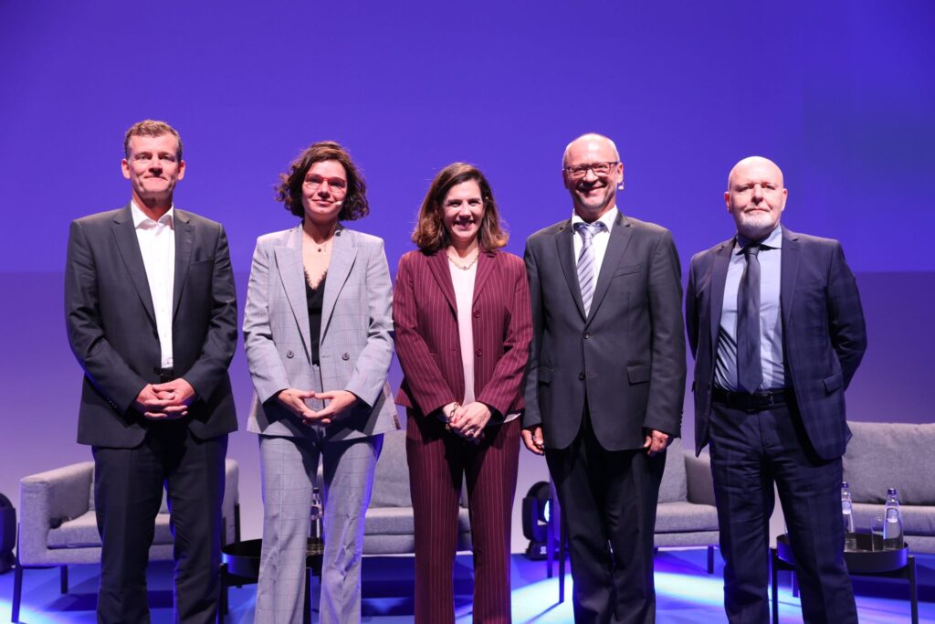 Five people, dressed in business attire, stand on a stage with purple lighting behind them. There is a woman in a gray suit, two men in dark suits, and two other individuals in lighter-colored suits, one man and one woman. They are posing and smiling at the camera.